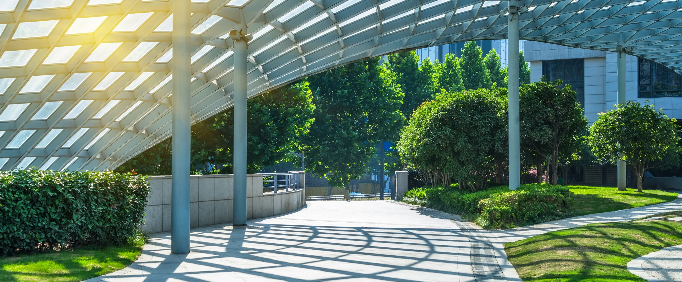 Curved glass canopy casting patterned shadows over a paved walkway surrounded by green bushes and trees in an urban park.