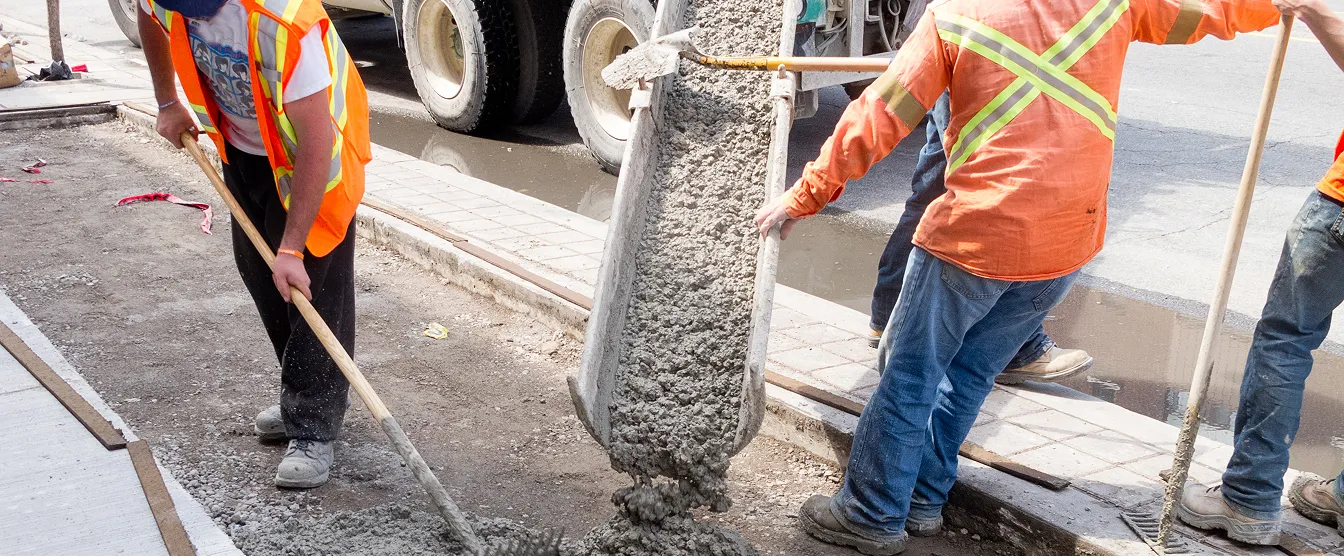 Construction workers in safety vests pouring and spreading concrete on a street sidewalk.