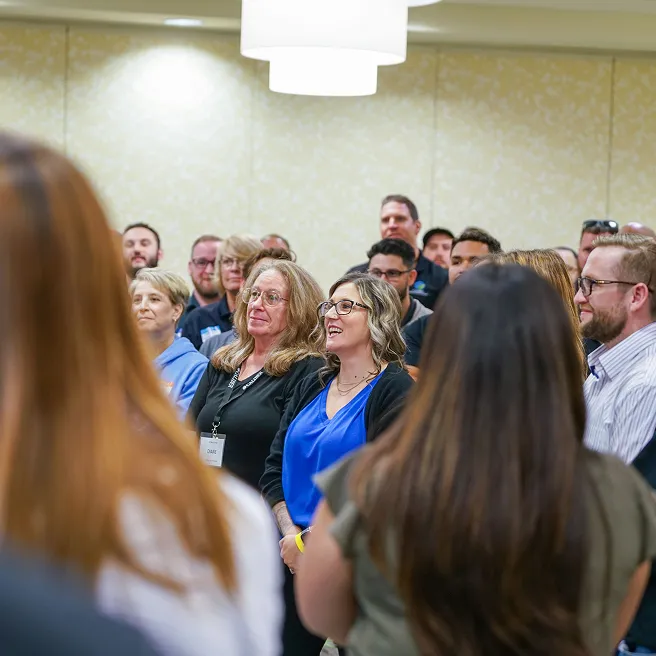 Group of 20 people smiling outdoors with a mountain range and cloudy sky in the background.