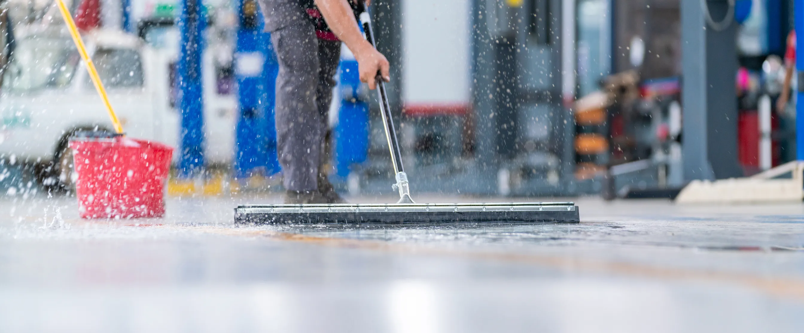 Person cleaning a wet floor with a floor squeegee and red bucket nearby in an indoor setting.