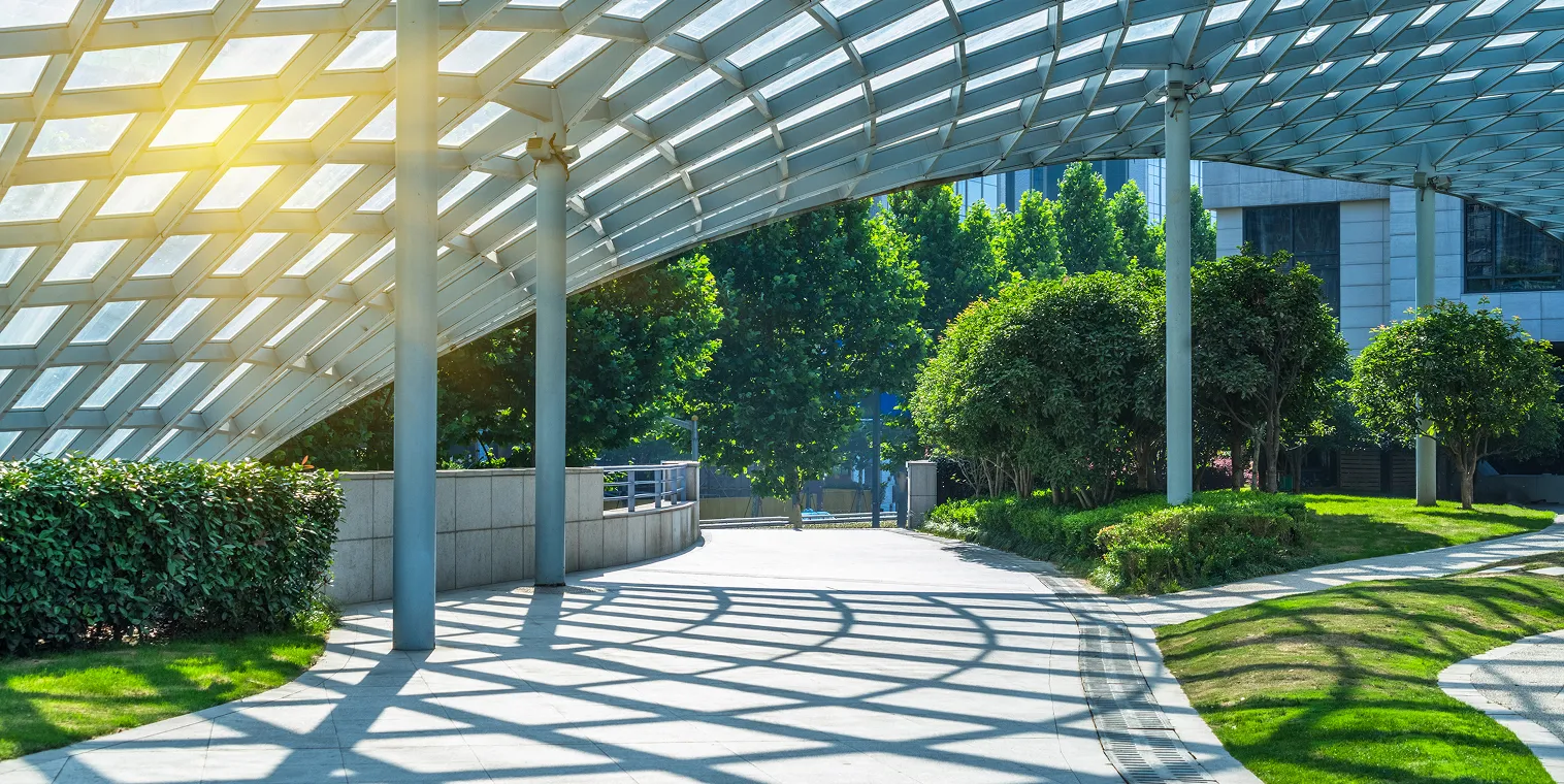 Sunlit modern walkway with a glass roof casting geometric shadows, surrounded by green trees and bushes.