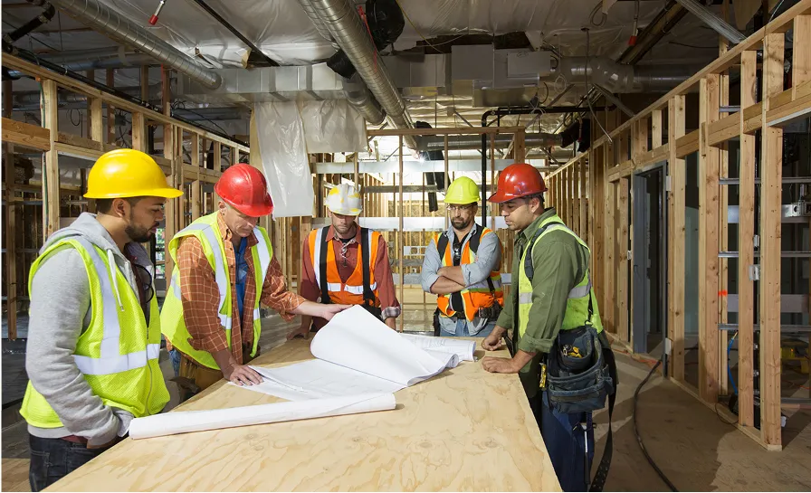 Five construction workers wearing safety helmets and vests examining blueprints on a wooden table inside a building under construction.
