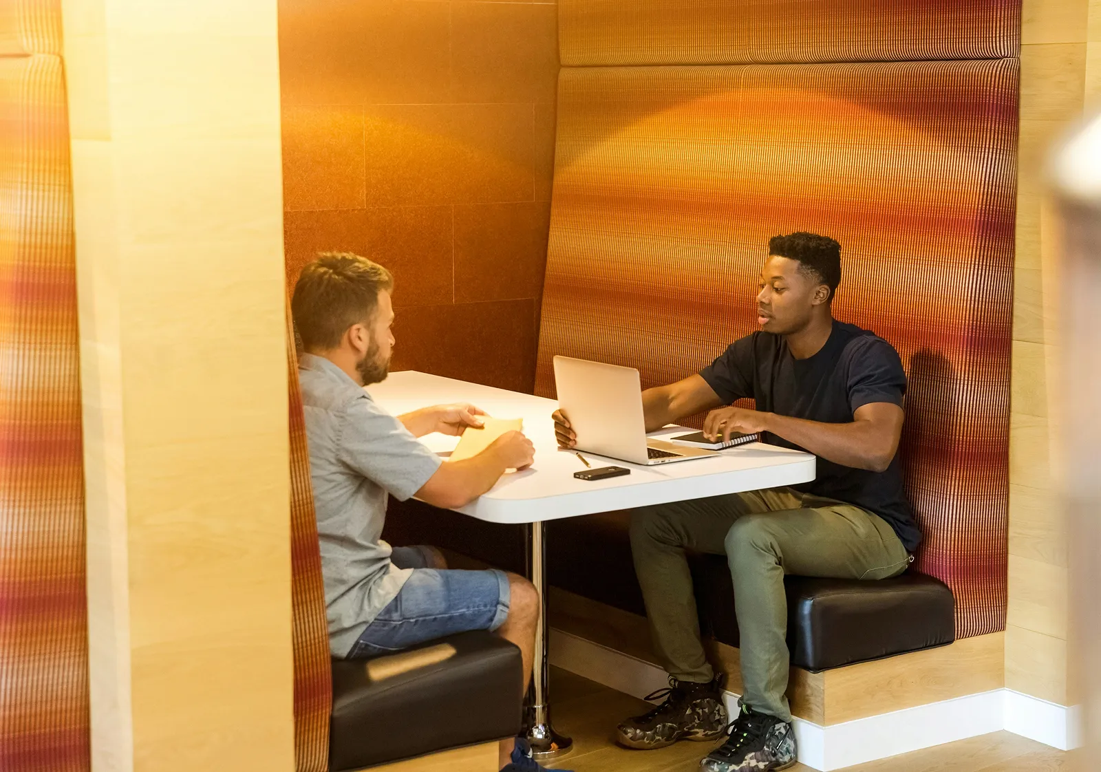 Two men sitting in a modern booth area, one using a laptop while the other holds a yellow folder.