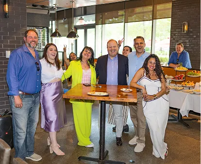 A group of eight people smiling and posing around a tall wooden table in a bright indoor setting with food trays in the background.