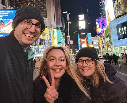Three smiling adults wearing winter jackets and hats taking a selfie at night in a brightly lit urban area with billboards and stores.