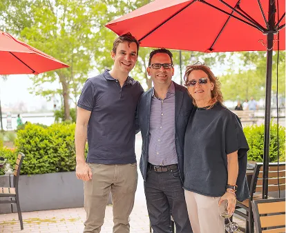 Three people standing together outdoors near red patio umbrellas, smiling at the camera.