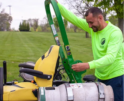 Man wearing a bright green long-sleeve shirt operating a green lawn mower in a grassy outdoor area.