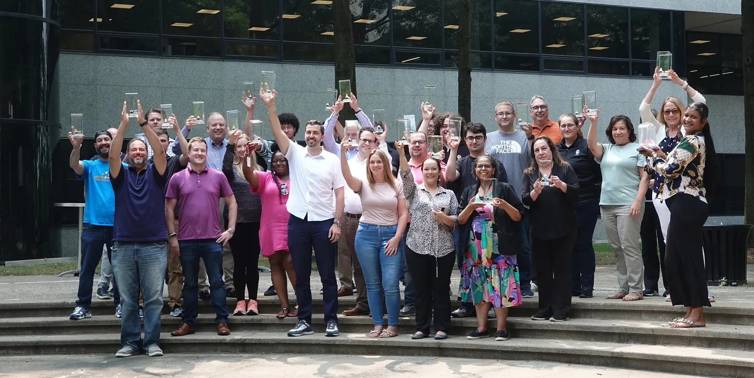Group of diverse people standing on steps outdoors holding up clear rectangular awards in celebration.