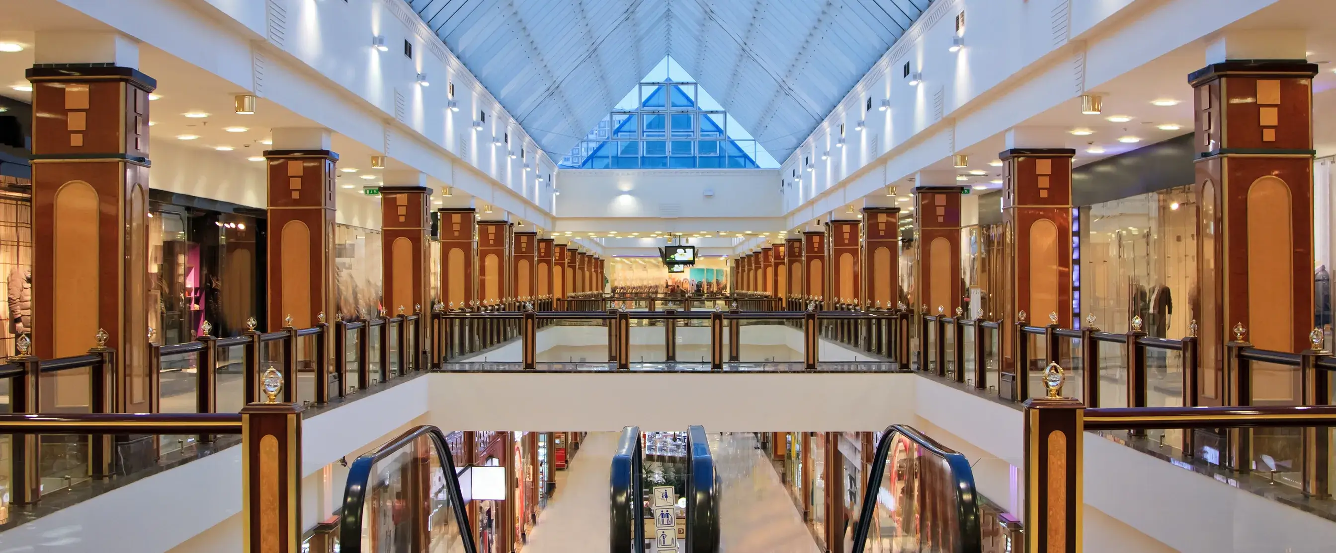 Interior of a spacious shopping mall with escalators, decorated wooden pillars, and a glass ceiling with a pyramid-shaped window.