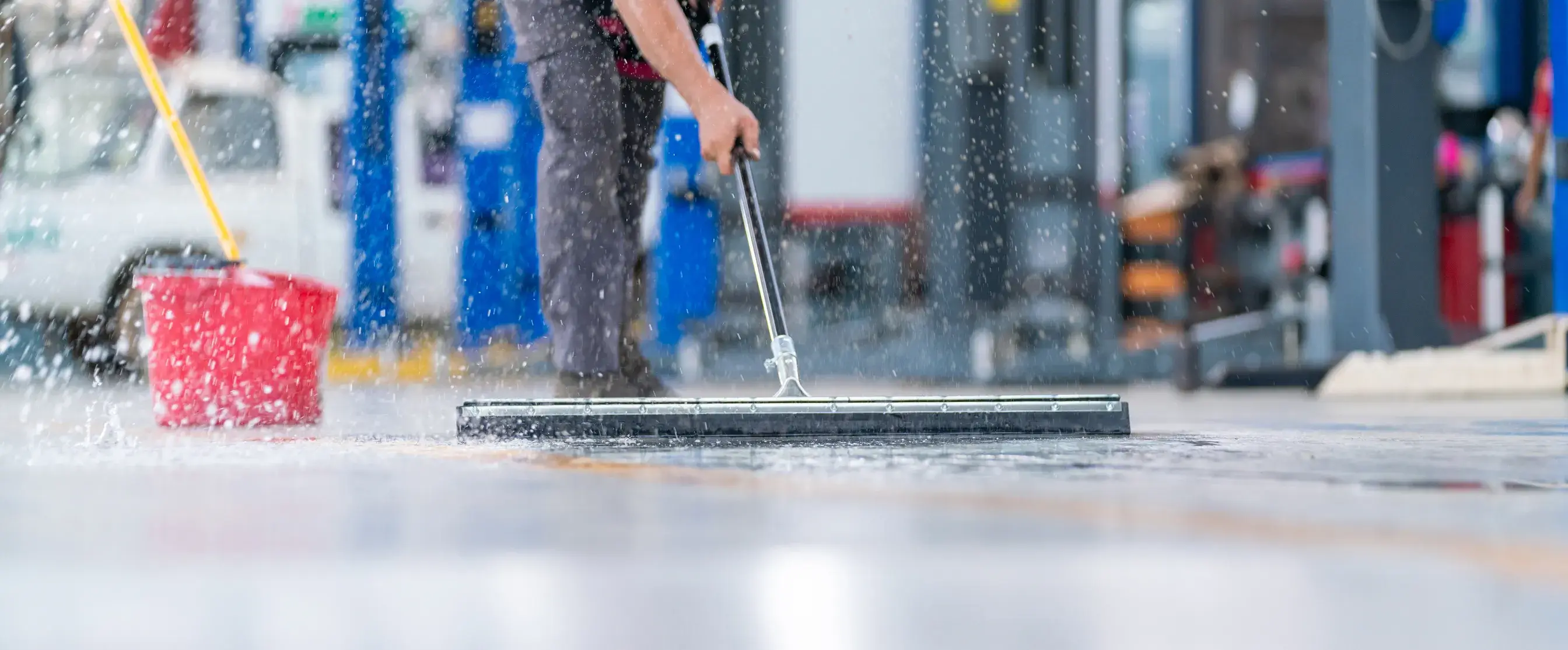 Person cleaning a wet floor with a floor squeegee and red bucket nearby in an indoor setting.