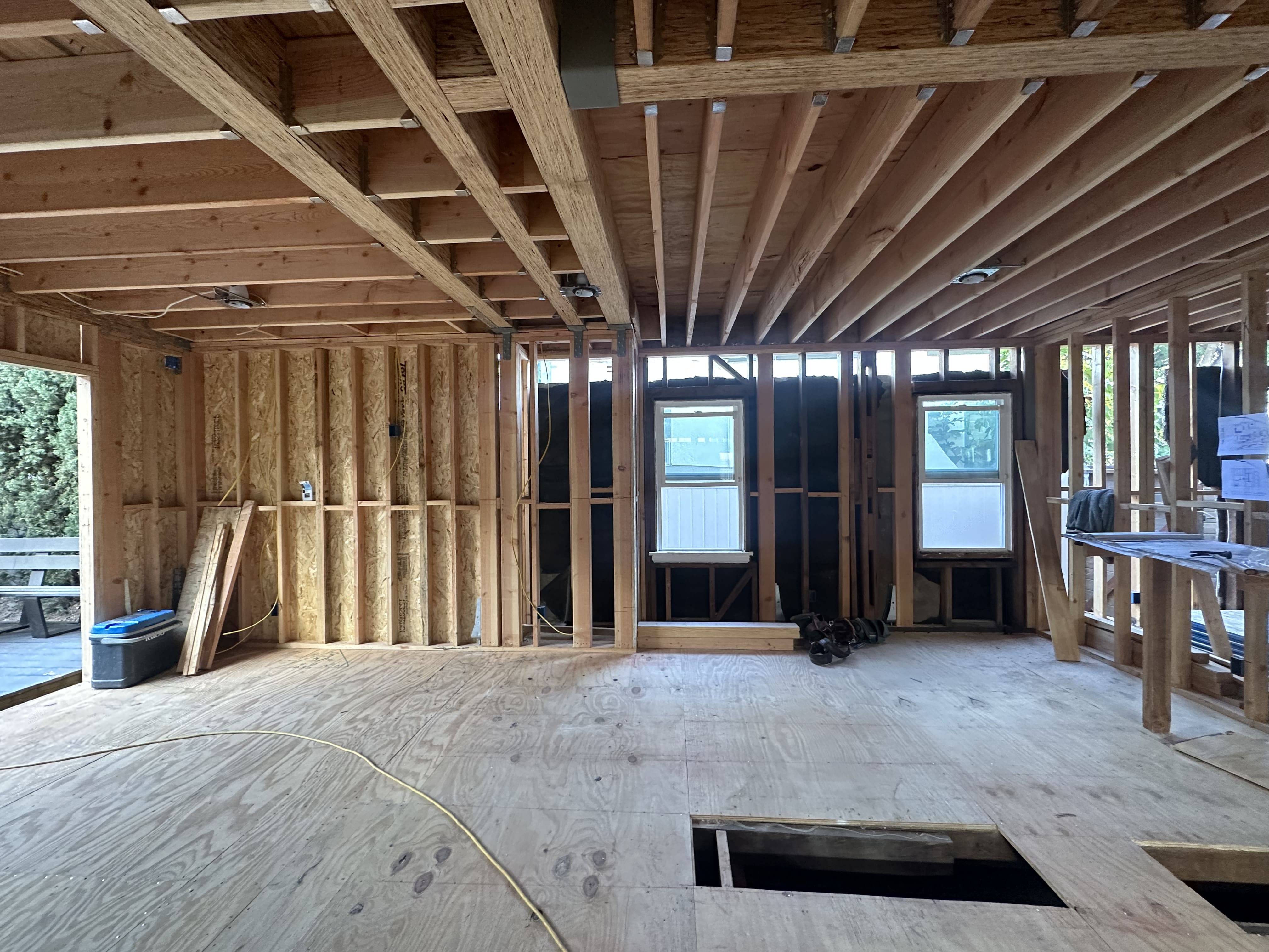 Interior of a wooden house under construction showing exposed framing, plywood floor, two windows, and construction tools.