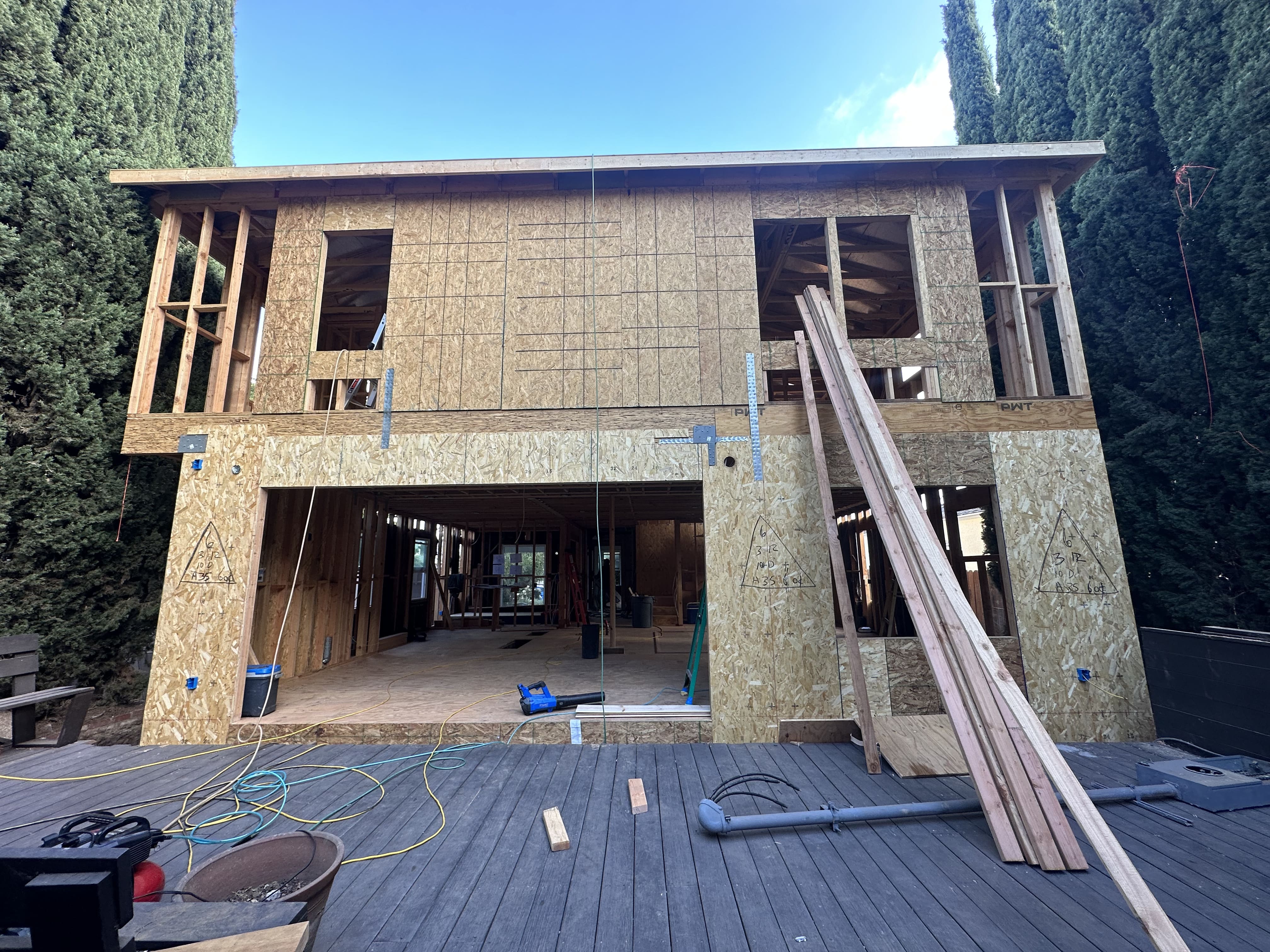 Two-story wooden house under construction with exposed framing, plywood sheathing, and lumber leaning against the front on a gray deck.