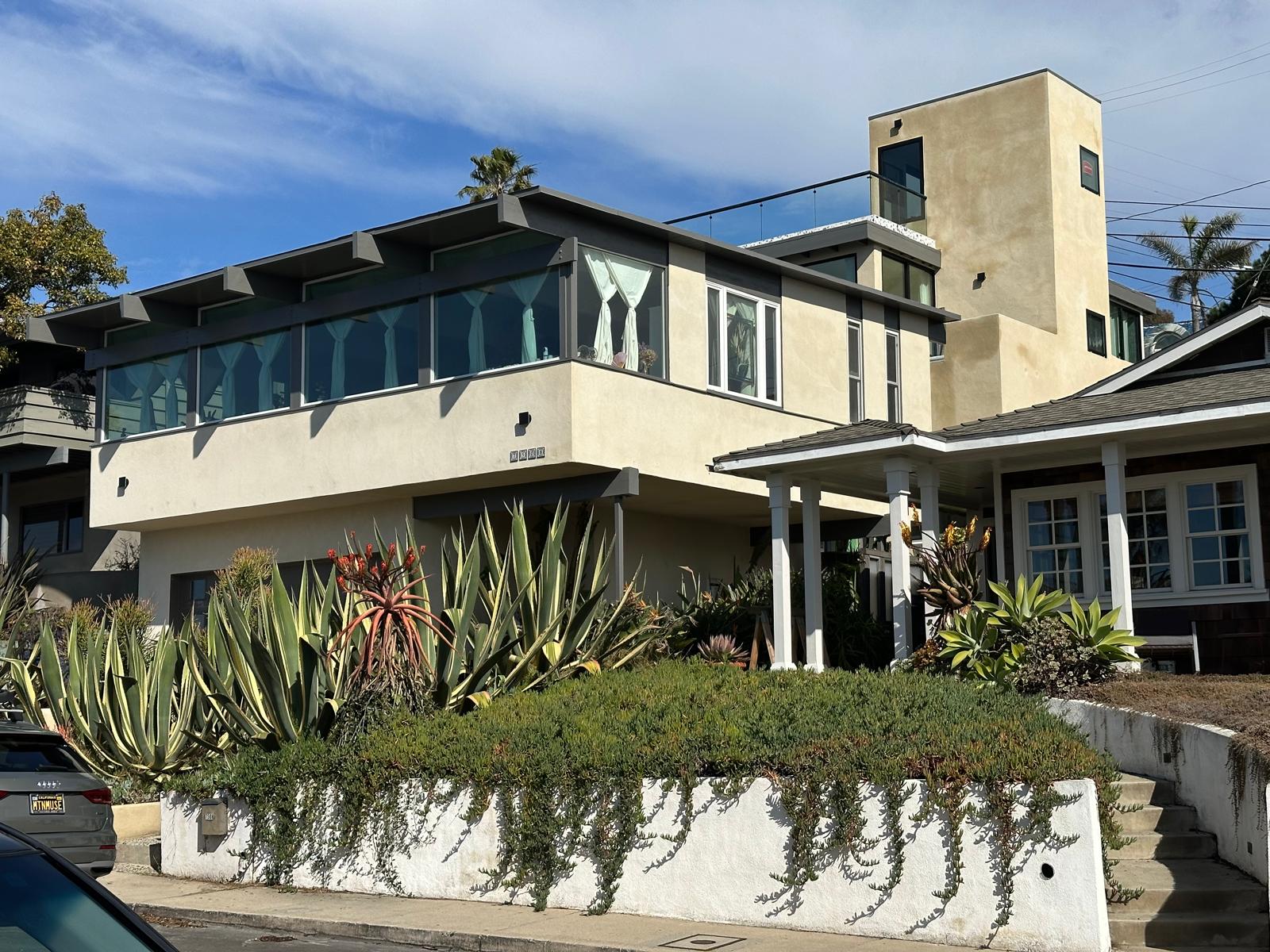 Modern multi-level house with large glass windows, beige exterior walls, and greenery in front garden under a blue sky.