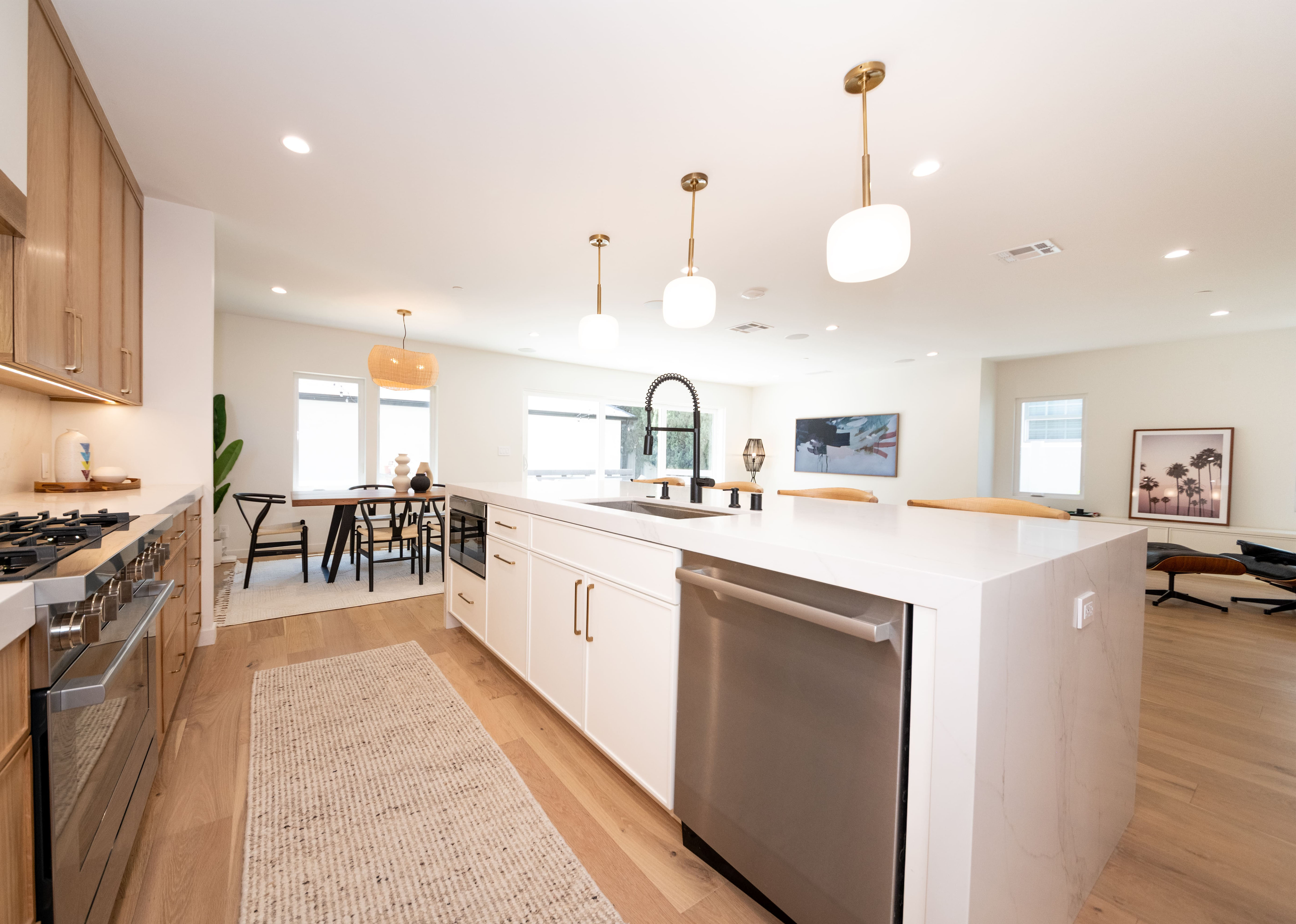 Modern open kitchen with white island, stainless steel appliances, wood cabinets, and dining area with pendant lights.