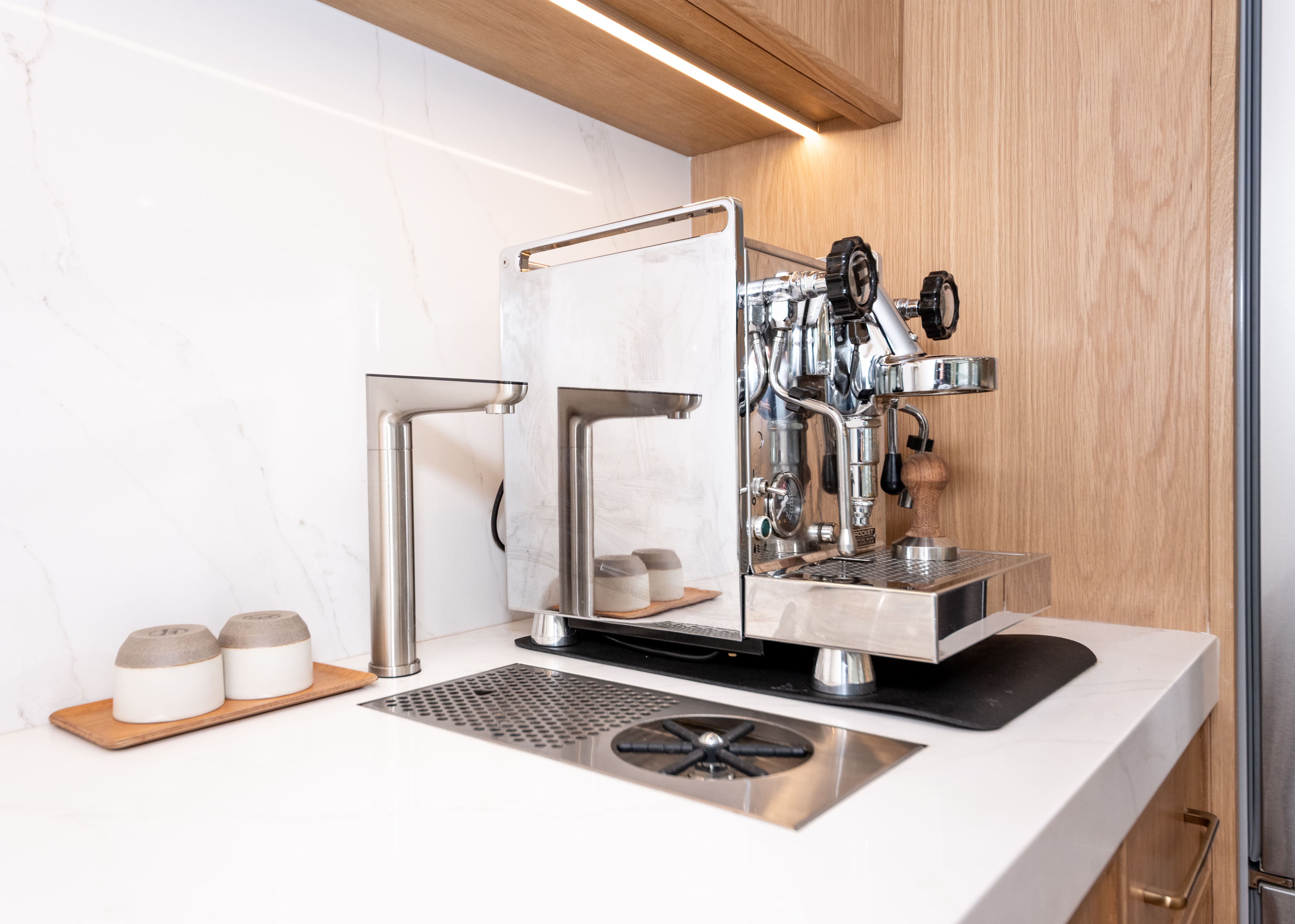 Modern espresso machine with wooden tamper on a white countertop next to two small containers on a tray and a built-in metal sink.