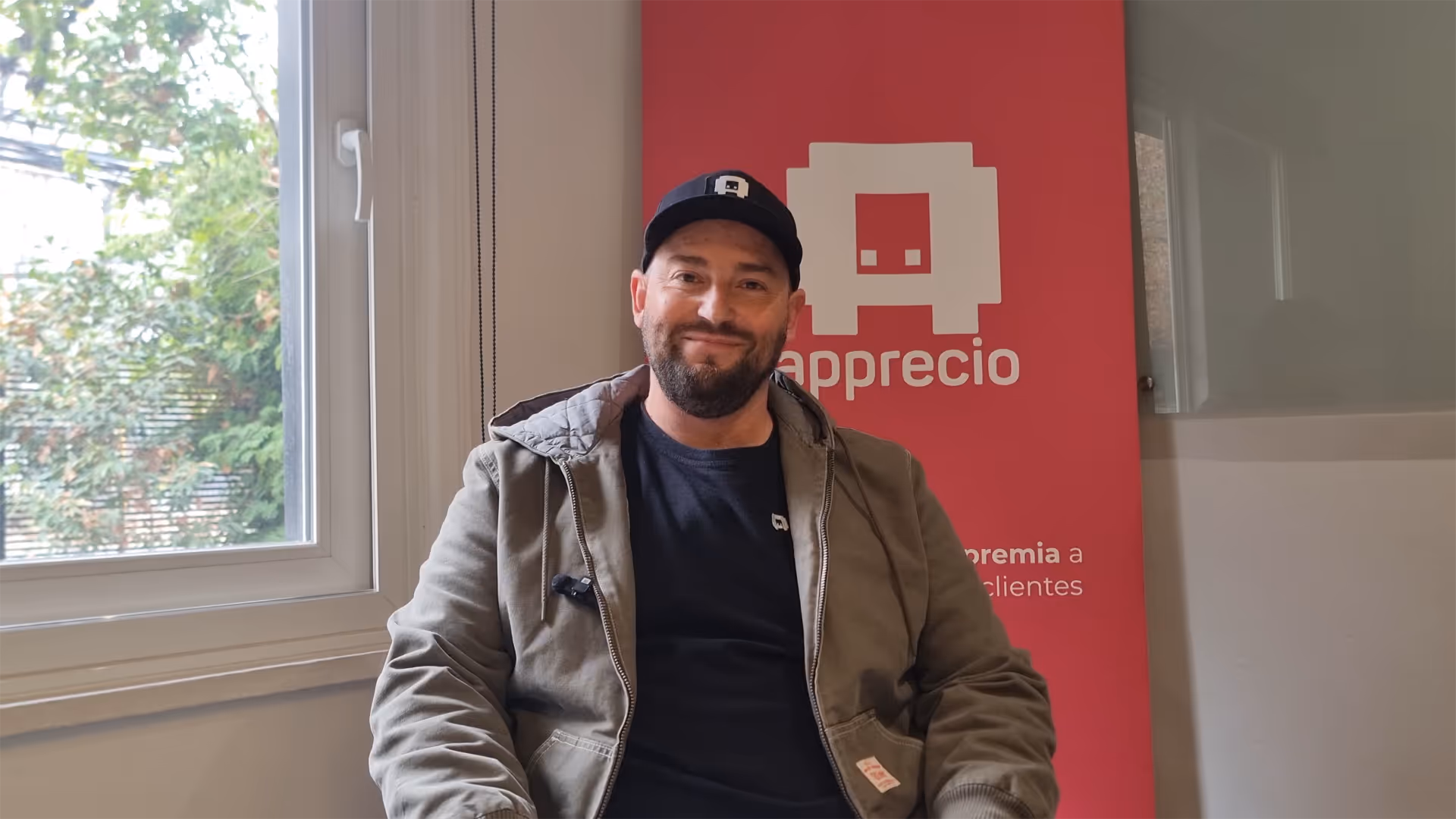 A man wearing a black hat and jacket sits in front of a red and white sign.
