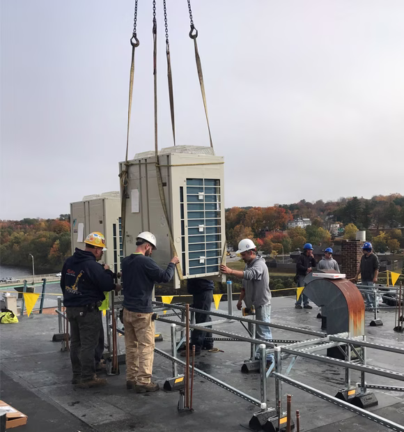 Construction crew installing large HVAC units on a rooftop using a crane, with workers guiding the equipment into place.