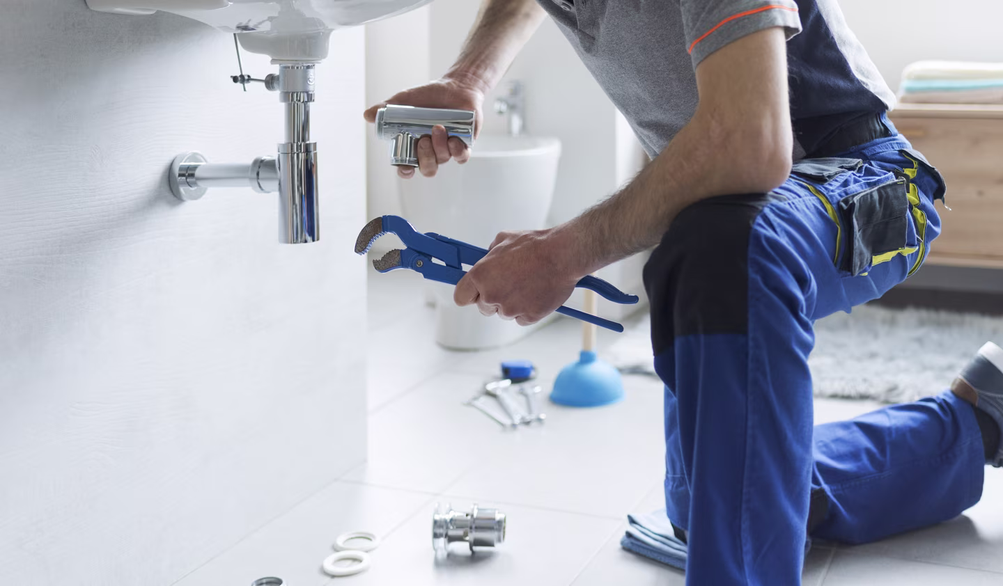 Plumber kneeling on the floor repairing bathroom sink pipes with tools spread around.