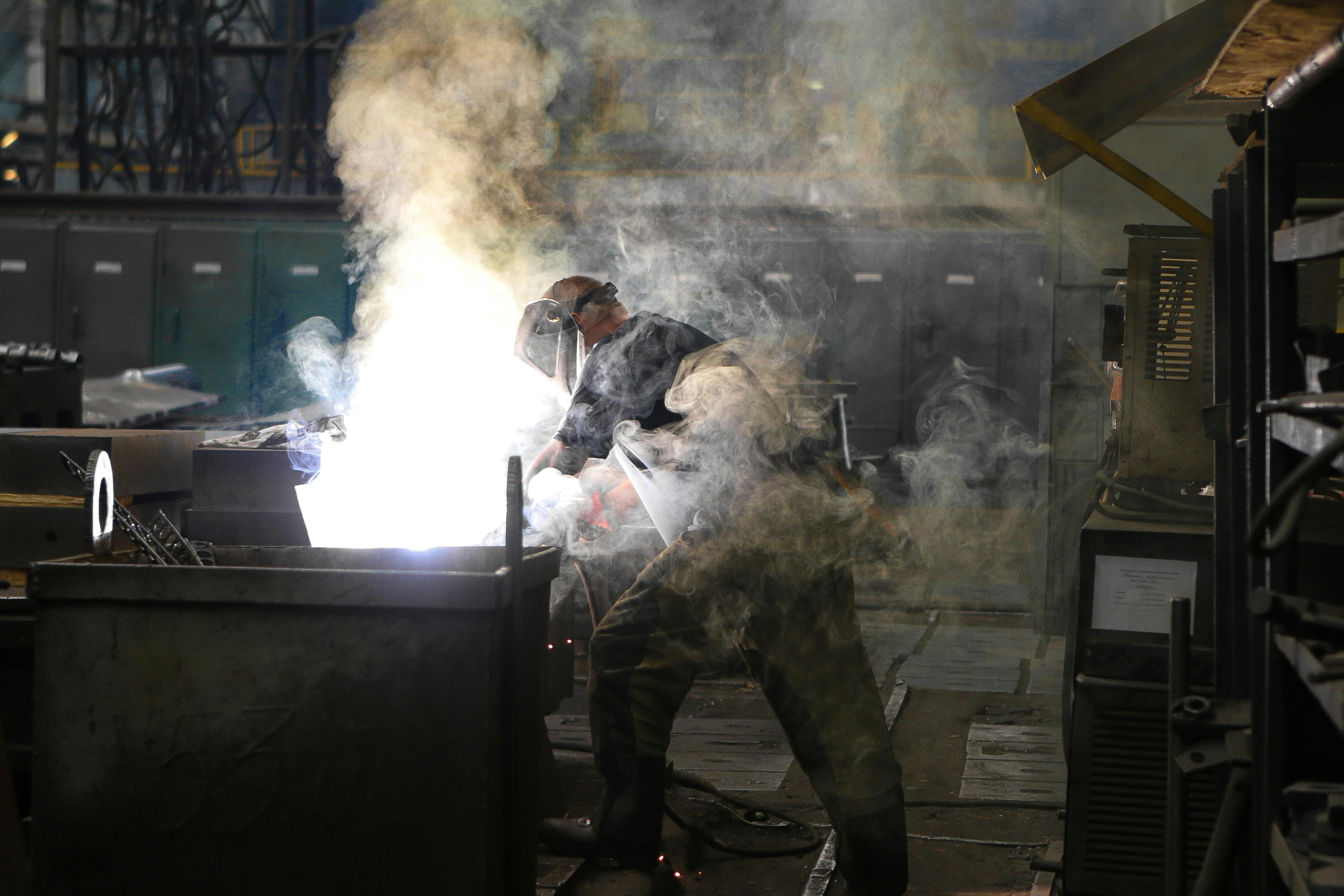 A worker wearing protective gear welding metal inside an industrial workshop with smoke surrounding him.