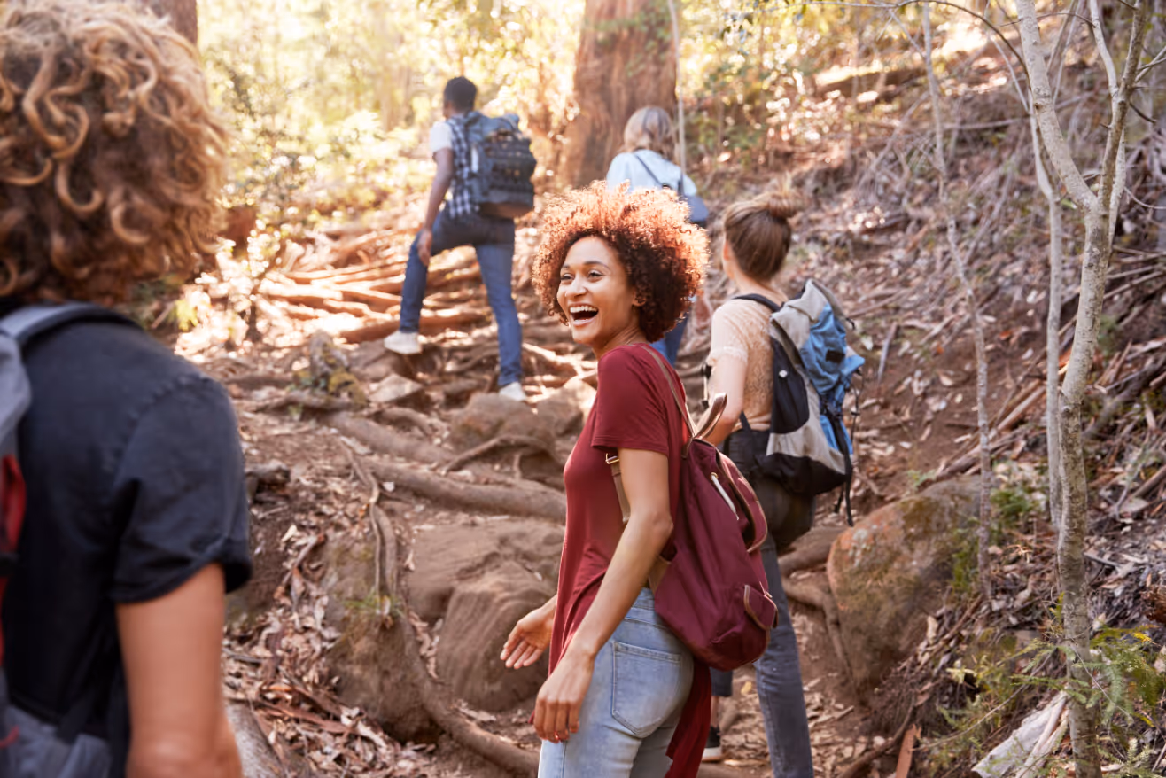 A group of young adults hiking on a wooded trail on a sunny day. One person is smiling back toward the camera, emphasizing nearby recreational opportunities and outdoor access.