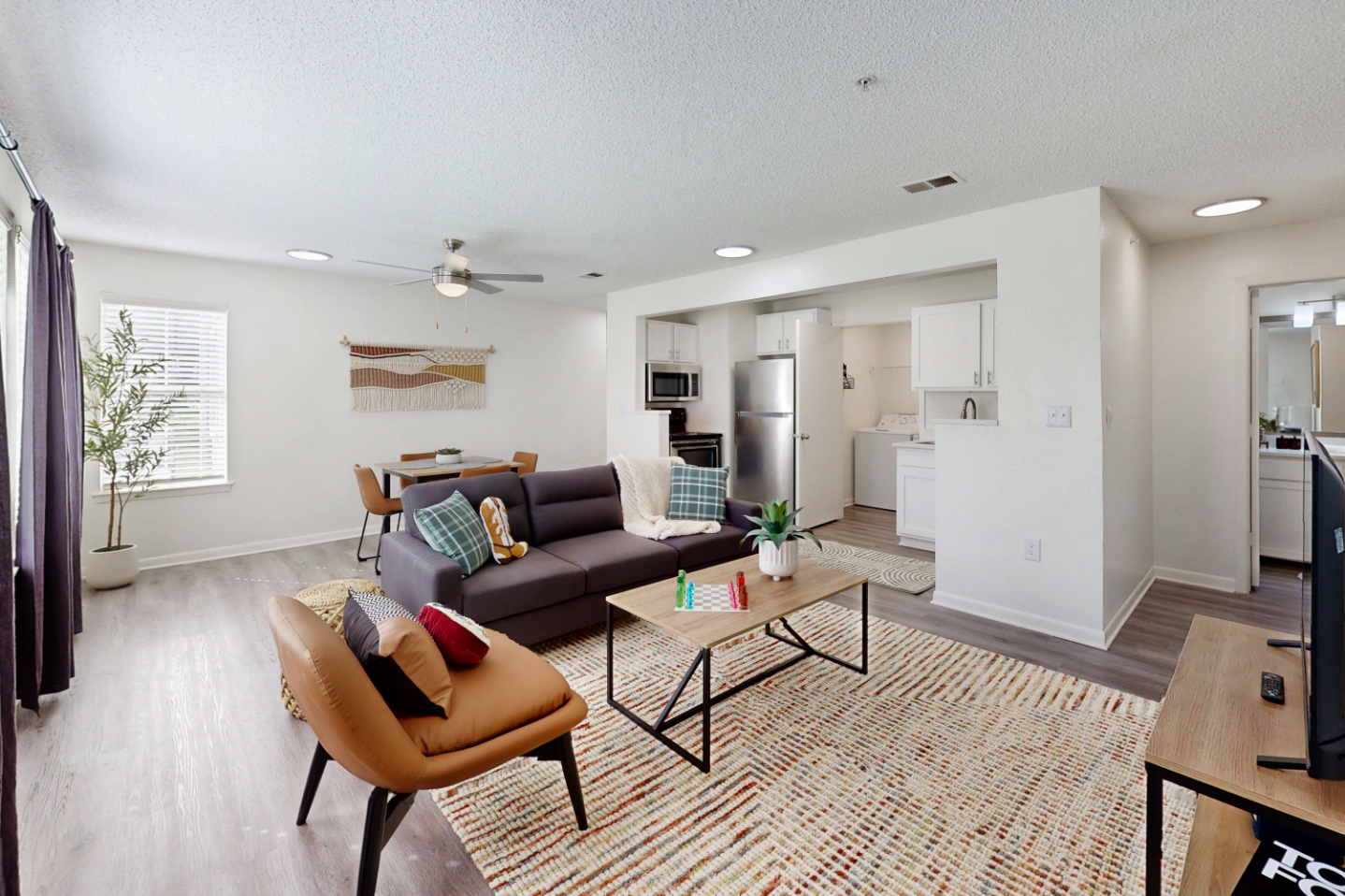 A wide view of a furnished apartment common area with a sofa and armchair on a white rug, looking toward a kitchen with a stainless steel refrigerator and laundry room