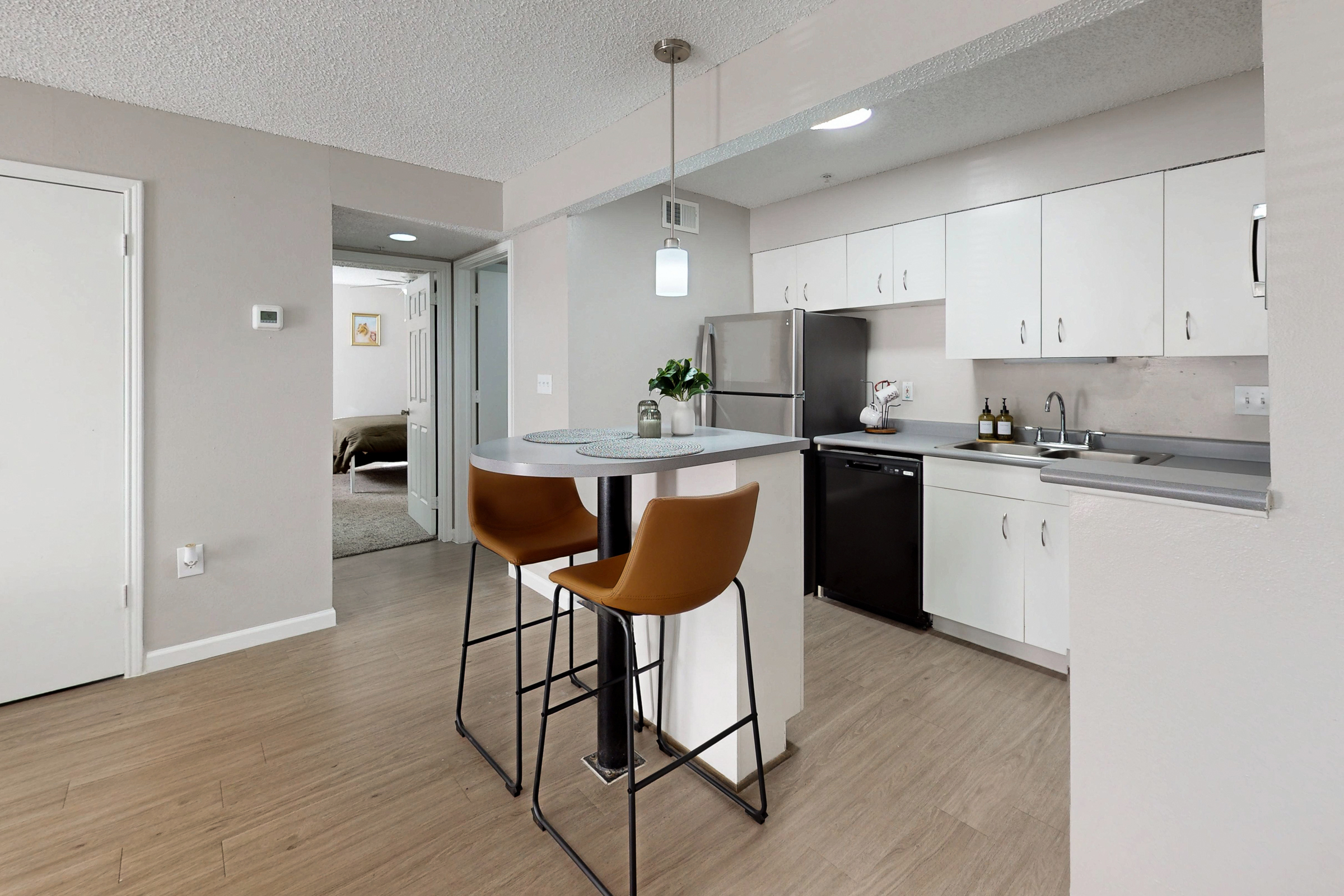 A wide view of a furnished apartment common area with a sofa and armchair on a white rug, looking toward a kitchen with a stainless steel refrigerator and laundry room