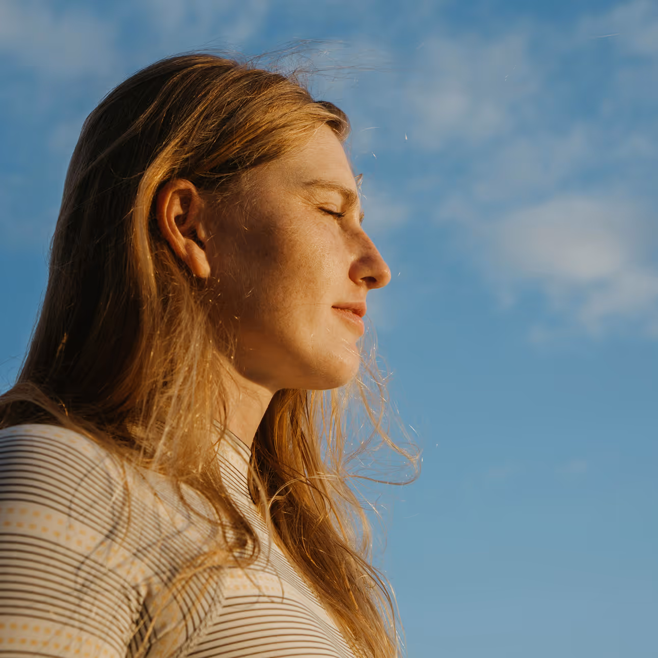 Mujer con ojos cerrados disfrutando del sol al aire libre con cielo azul de fondo.