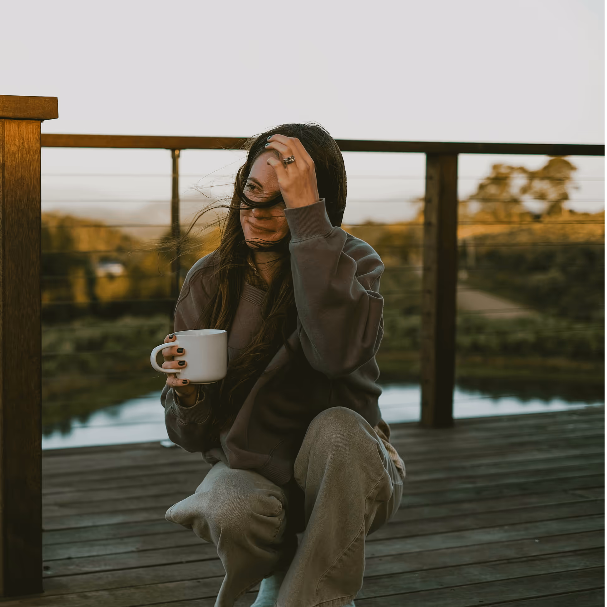 Mujer con suéter gris y pantalones cómodos en un balcón de madera sosteniendo una taza blanca mientras el viento mueve su cabello.