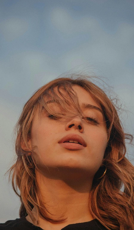 Primer plano de una mujer joven con cabello castaño claro y suelto mirando hacia abajo con el cielo azul de fondo.