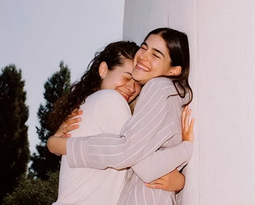 Dos mujeres sonrientes abrazándose con ojos cerrados frente a una pared blanca y árboles al fondo.