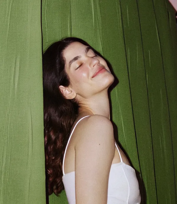 Mujer con cabello castaño rizado y camiseta blanca de tirantes, apoyada contra unas cortinas verdes con expresión de tranquilidad y ojos cerrados.