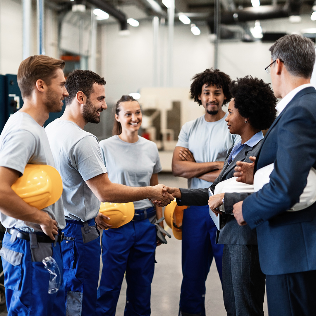 Group of construction workers and business professionals shaking hands in a factory setting, symbolizing successful partnership and collaboration.