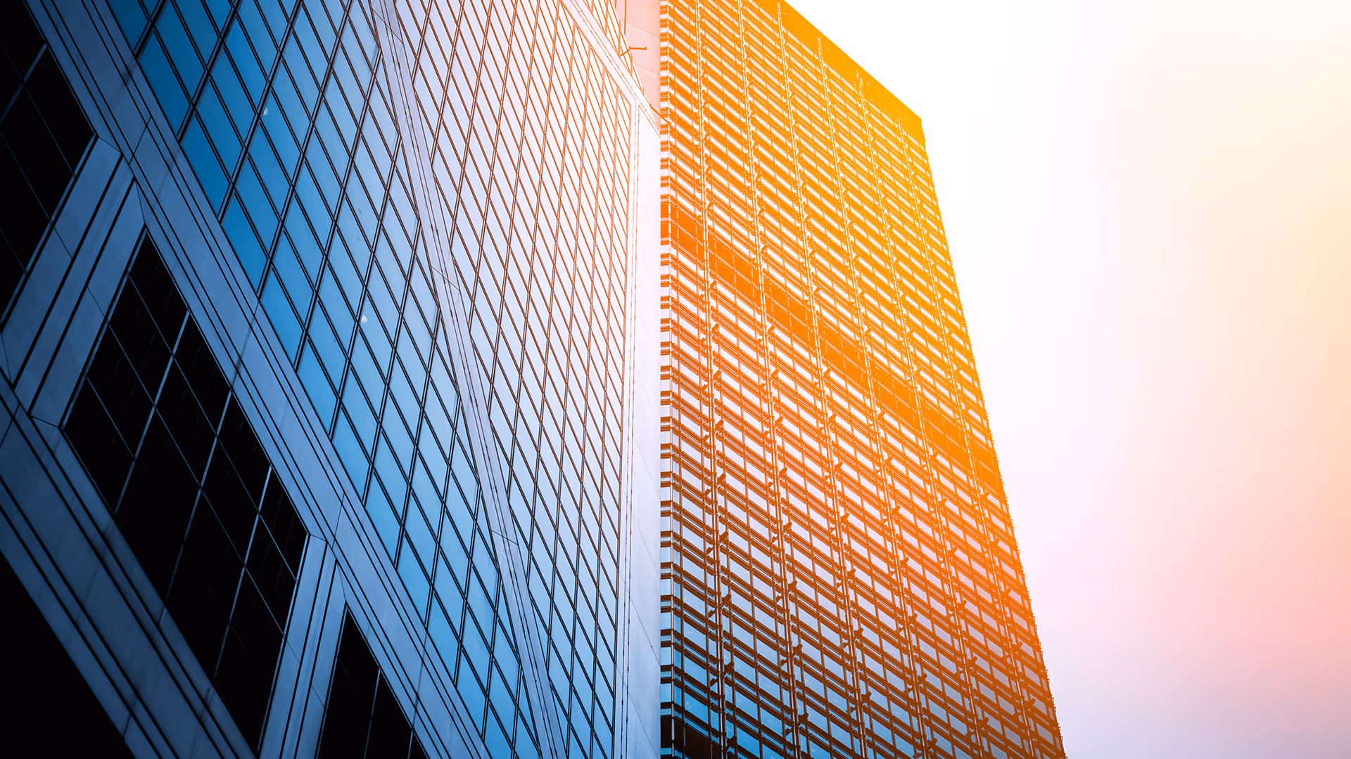 Upward view of two modern skyscrapers with reflective glass windows, one illuminated by orange sunlight.