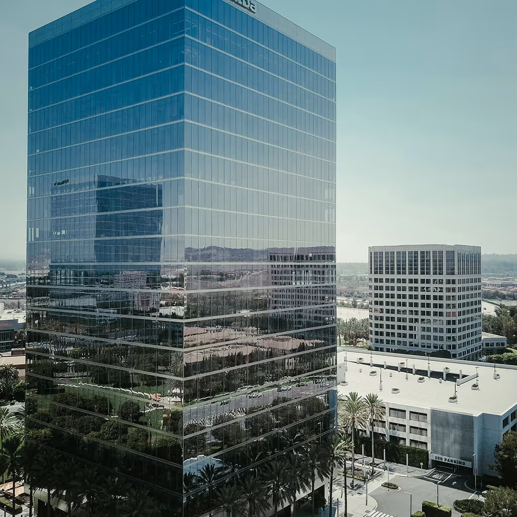 Tall modern glass office building reflecting its surroundings, with palm trees and adjacent parking structures under a clear sky.