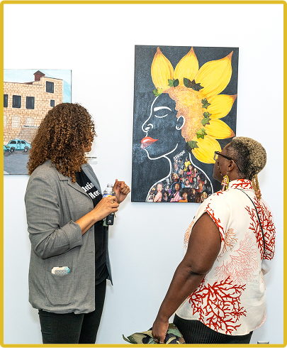 Two women viewing colorful flower artwork at a gallery; one holds a drink.