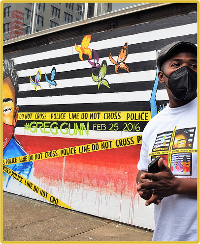 Man wearing a mask stands near colorful mural with butterfly designs and crime scene tape, conveying a social message.