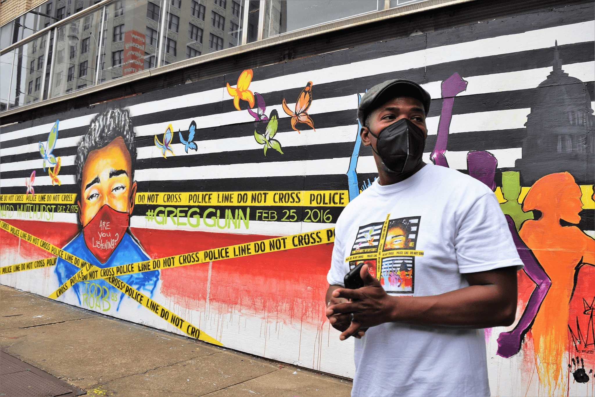 Man in mask stands by colorful mural with bold graphics and portraits, wearing a t-shirt, in an urban setting.