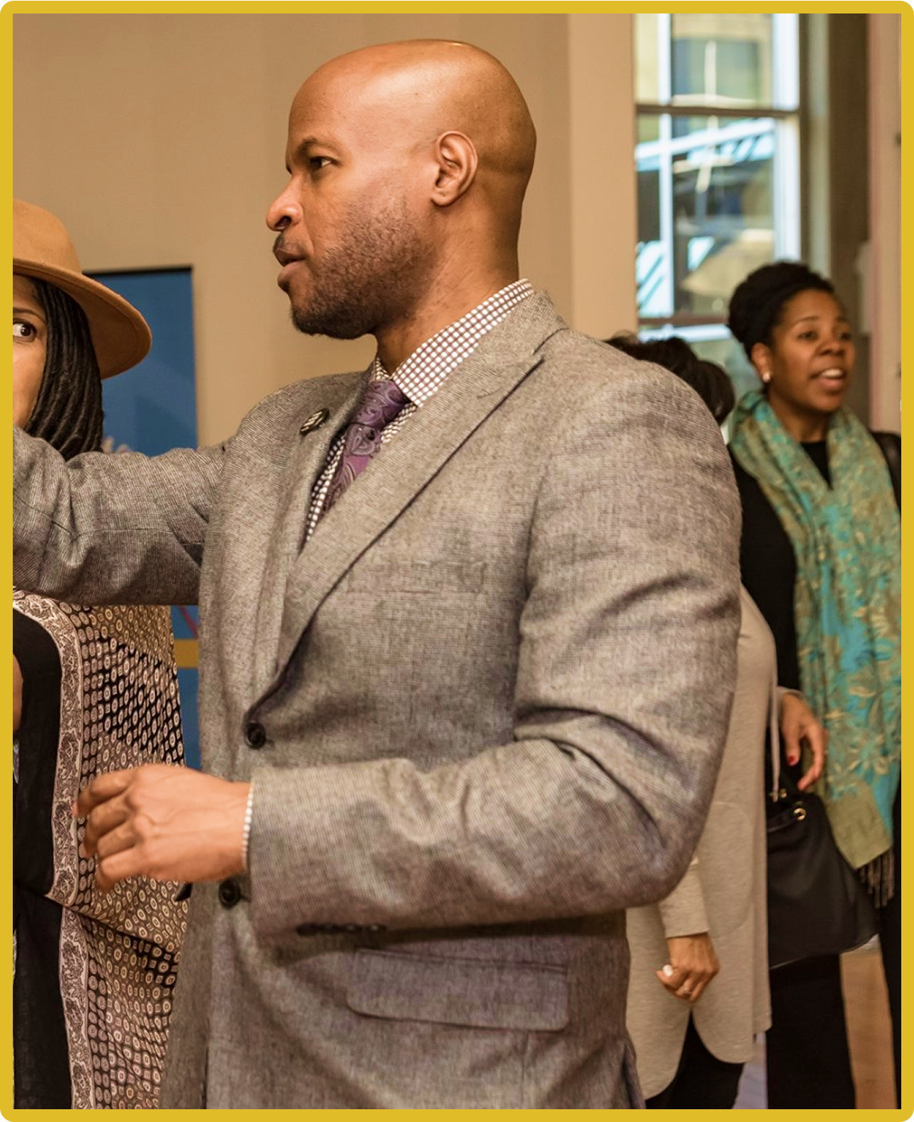 A man in a gray suit gestures, with people in the background. Indoors, social setting.