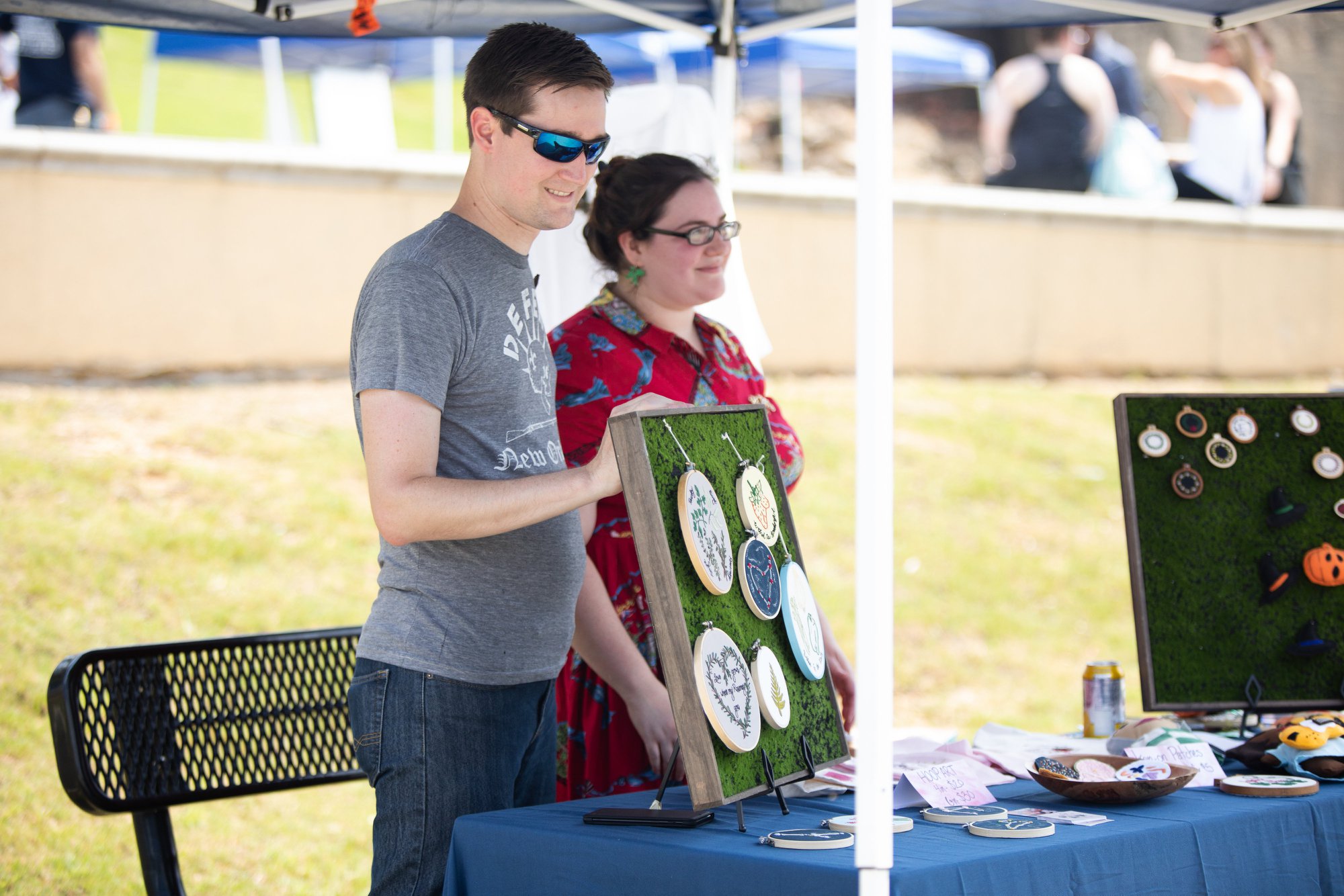 People stand at a table outdoors displaying art or crafts on a sunny day.