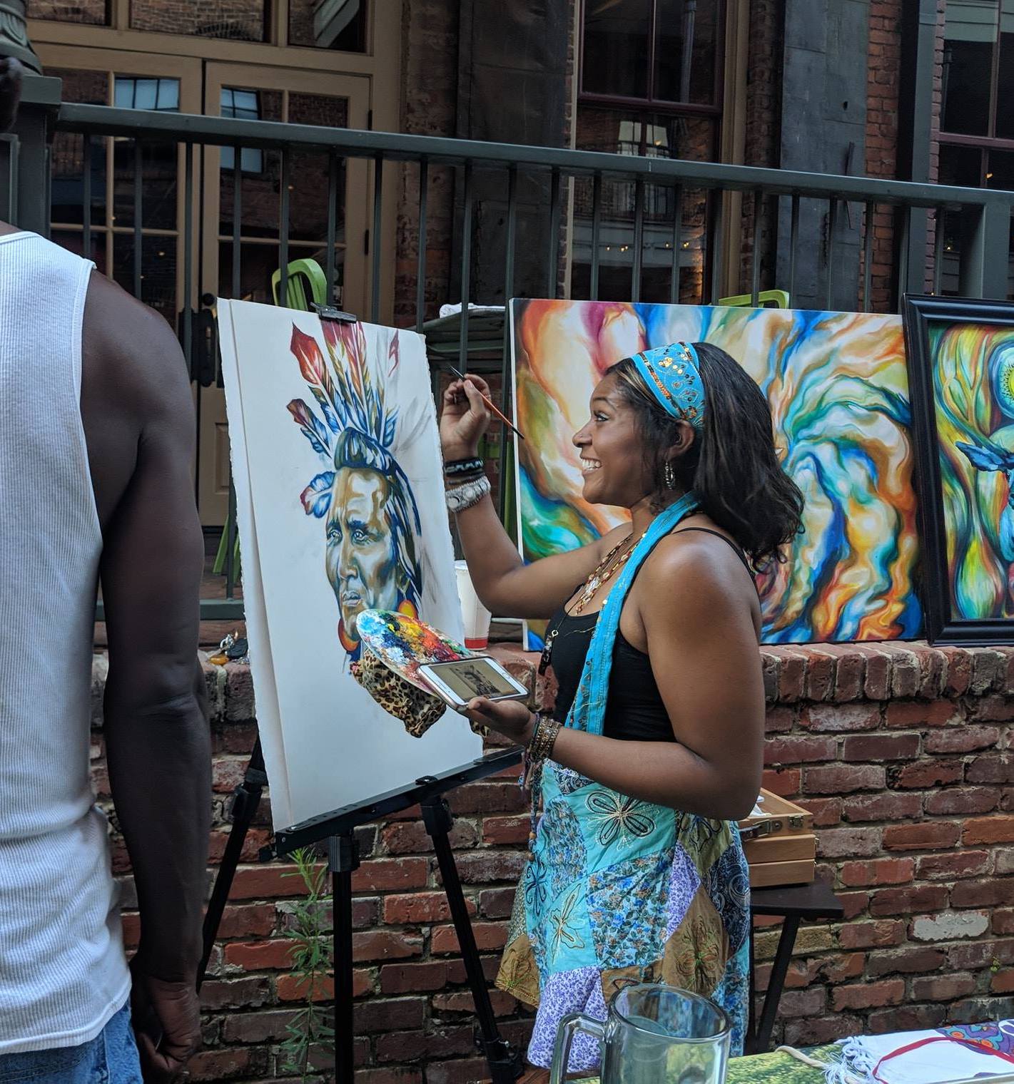 Artist painting outdoors with vibrant artwork in the background.