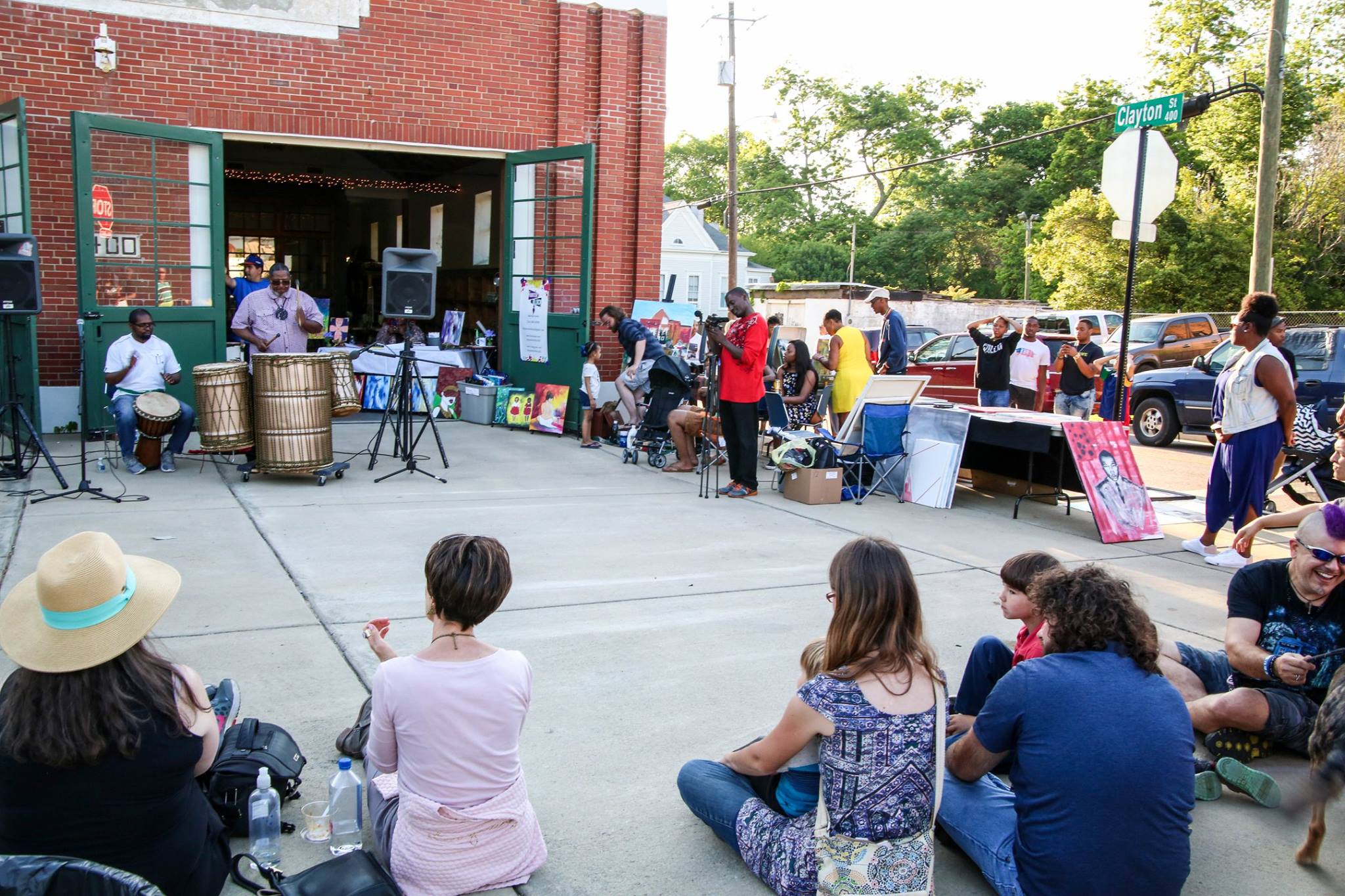Outdoor community event with people gathering, sitting on the ground, and listening to a speaker near a building.