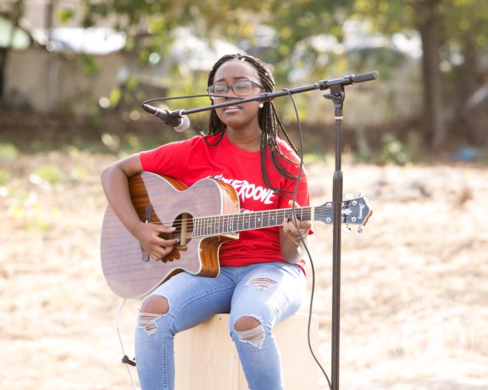 Person playing guitar outdoors, wearing a red shirt, seated with a microphone.