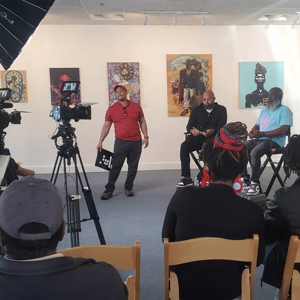 Panel discussion filmed in gallery, audience seated, cameras recording, portraits on walls, casual setting.