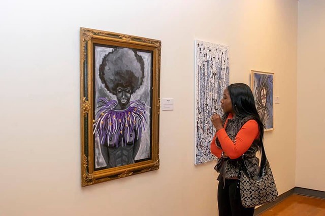 Man in mask stands by colorful mural with bold graphics and portraits, wearing a t-shirt, in an urban setting.