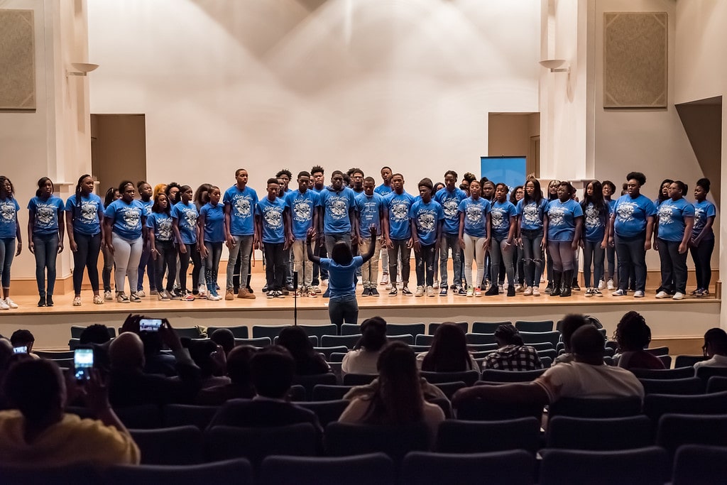 People stand at a table outdoors displaying art or crafts on a sunny day.
