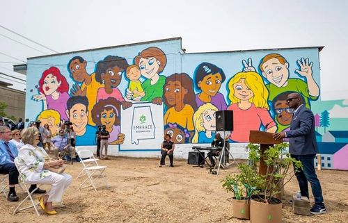 Person playing guitar outdoors, wearing a red shirt, seated with a microphone.