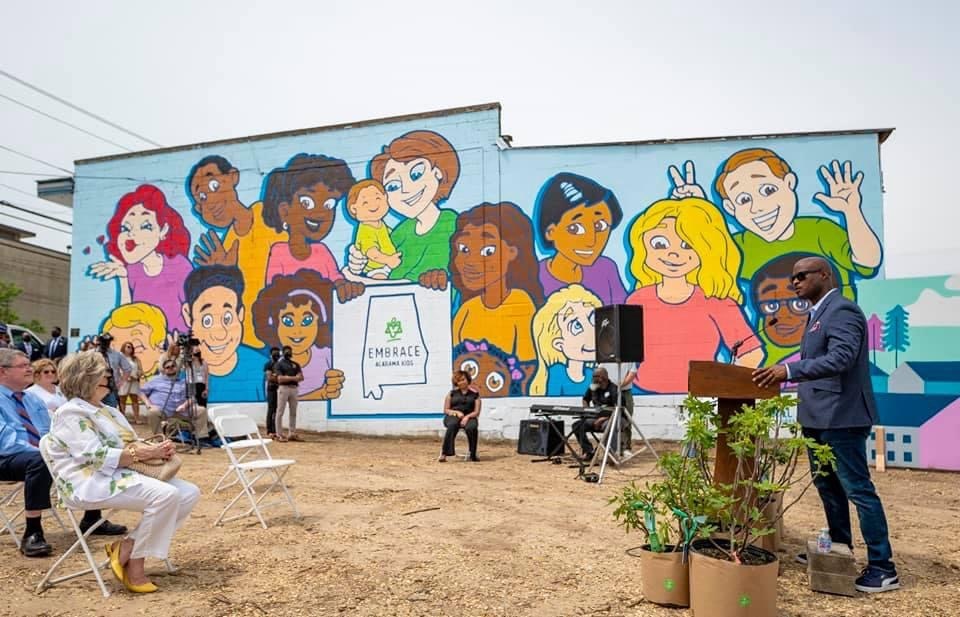 Person playing guitar outdoors, wearing a red shirt, seated with a microphone.