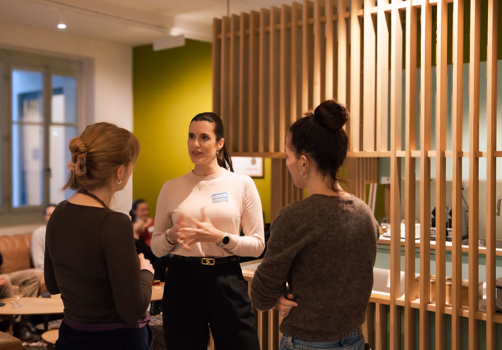 Three women engaged in conversation in a modern office lounge with wooden slats and a green accent wall.