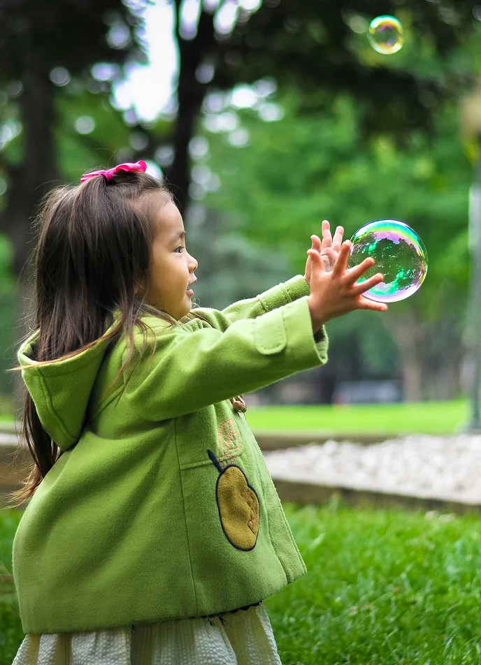 Young girl in a green coat joyfully reaching out to catch a soap bubble in a park.