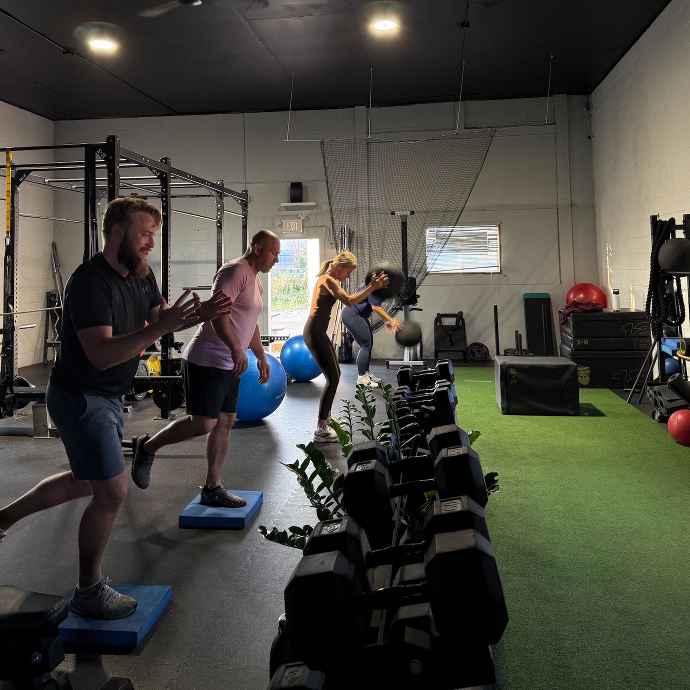 Three people exercising inside a gym using balance pads and medicine balls with dumbbells and fitness equipment nearby.